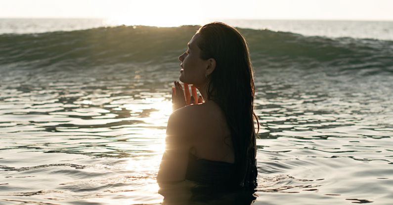 Healthy Travel - Side view of tranquil young female tourist with long dark hair standing in waving ocean with closed eyes and enjoying summer sunset