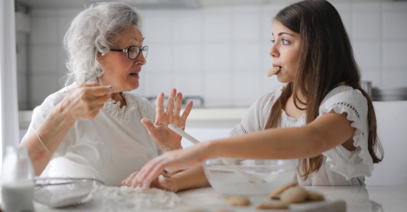 Cooking Kids - Calm senior woman and teenage girl in casual clothes looking at each other and talking while eating cookies and cooking pastry in contemporary kitchen at home