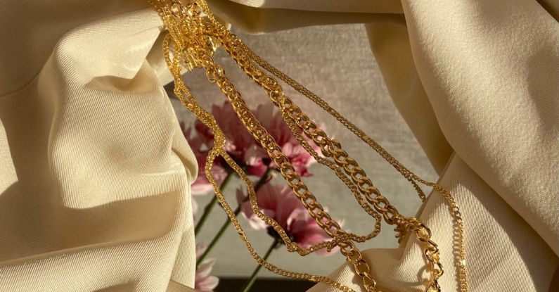 Fabric Decor - From above of golden chain with pendant placed on crumpled fabric with mirror near pink flowers in light stylish room