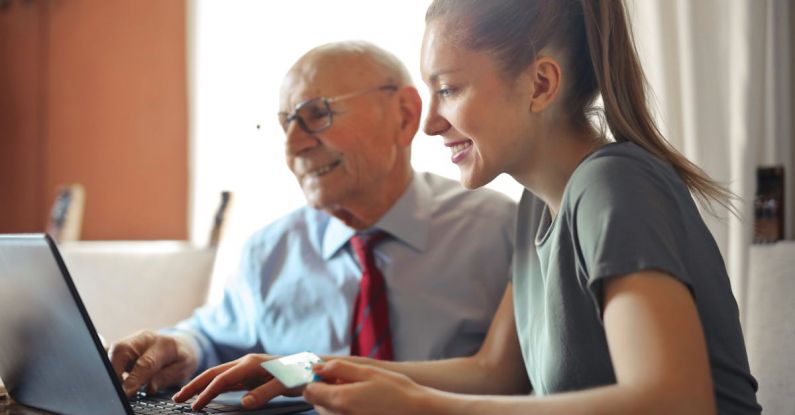 Family Finance - Young woman in casual clothes helping senior man in formal shirt with paying credit card in Internet using laptop while sitting at table